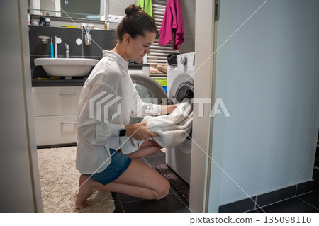 A woman loads towels into a washing machine in a home bathroom. Real life interior scene showing everyday laundry routine, household responsibility and domestic work. A woman loads towels into a washing machine in a home bathroom. Real life interior scene showing everyday laundry routine, household responsibility and domestic work. 135098110
