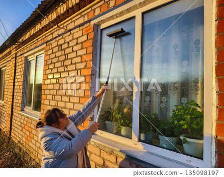 A woman goes about her housework, washing the outside window of a brick house with a scrub brush in the warm evening sun. During the golden hour, the woman washes the outside window of the house. 135098197