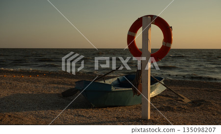 An orange life preserver hangs from a wooden pole next to a turquoise rowboat on a pebble beach. Calm sea waves roll under a serene sunset sky, symbolizing themes of safety, rescue, and tranquility. An orange life preserver hangs from a wooden pole next to a turquoise rowboat on a pebble beach. Calm sea waves roll under a serene sunset sky, symbolizing themes of safety, rescue, and tranquility. 135098207