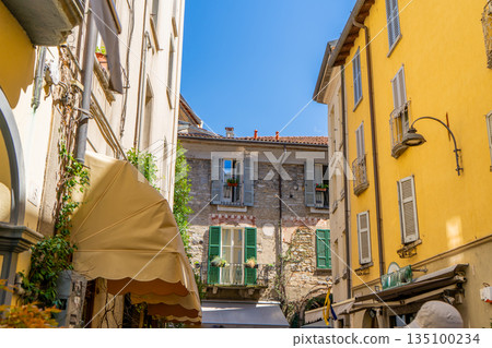 Multicolor windows balcony with shutters. Italy empty narrow street in summer day. Italian style facade architecture. Yellow wall Multicolor windows balcony with shutters. Italy empty narrow street in summer day. Italian style facade architecture. Yellow wall 135100234