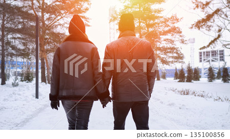 Couple Holding Hands Walking Through Snowy Park, Sunset Backlight Creates Warm Glow, Tall Pines Line Path, Bundled Jackets And Beanies, Peaceful Winter Atmosphere, Intimate Connection And Gentle Pace 135100856