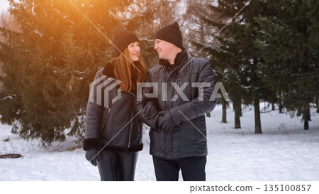 Couple Laughing Walking Near Evergreen Trees, Matching Beanies And Jackets, Playful Eye Contact And Shared Jokes, Soft Sunlight Filtering Through Branches, Candid Joyful Moment Captured Outdoors 135100857