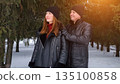 Couple In Winter Coats Looking Up, Man Hugging Woman Under Evergreen Trees With Warm Sunlight Flare, Beanies And Leather Jackets, Cozy Atmosphere, Candid Weekend Escape And Genuine Connection 135100858