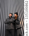 Fashionforward Pair In Dark Clothing Poses Near Architectural Background, Two Individuals Donning Black Jackets Stand Confidently Against Contemporary Striped Wall Showcasing Urban Style 135100862