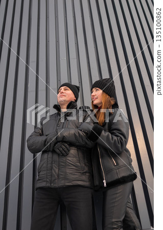 Fashionforward Pair In Dark Clothing Poses Near Architectural Background, Two Individuals Donning Black Jackets Stand Confidently Against Contemporary Striped Wall Showcasing Urban Style 135100862