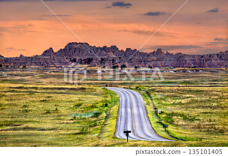 Badlands National Park landscape stands in South Dakota, USA. Scenic view features the winding Loop Road leading towards rugged rock formations and green prairie under a colorful sunset sky Badlands National Park landscape stands in South Dakota, USA. Scenic view features the winding Loop Road leading towards rugged rock formations and green prairie under a colorful sunset sky 135101405