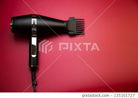 Close-up of a black hair dryer with a comb attachment to create volume on a pink background with space for text. The concept of long-term hair styling, hairstyles, curls and hair care. High quality Close-up of a black hair dryer with a comb attachment to create volume on a pink background with space for text. The concept of long-term hair styling, hairstyles, curls and hair care. High quality 135101727
