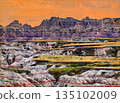 Big Badlands Overlook in Badlands National Park, South Dakota, USA. Scenic view features colorful eroded rock formations and grassy layers under a bright orange sunset sky 135102009
