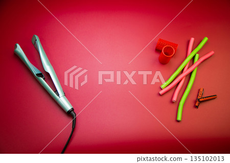 Top view of a green hair straightener with a swirling ceramic canvas, next to which there are curlers of different shapes and sizes on a pink background with space for text. The concept of styling Top view of a green hair straightener with a swirling ceramic canvas, next to which there are curlers of different shapes and sizes on a pink background with space for text. The concept of styling 135102013