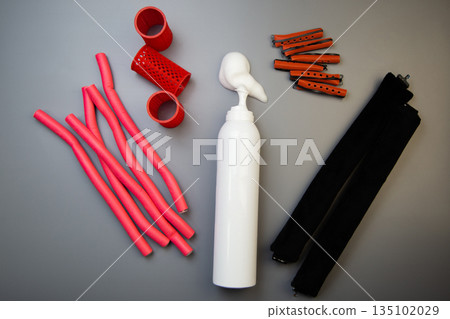 A close-up on a gray background shows a bottle of foam for long-term hair styling, white foam poured out of it, next to it there are curlers of different shapes and sizes. The concept of styling 135102029