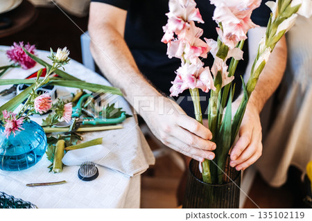 Hands hold large pink and white gladiolus stems above work table covered in botanical trimmings and floral tools. DIY floral design, creative skill-building, hobbyist floriculture. 135102119