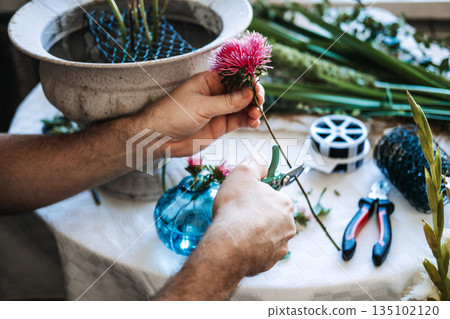 Hands use small snips to trim pink flower stem on table near stone urn and floral wire. Mindful floral arranging, botanical meditation, sensory hobbies. 135102120
