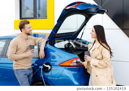 Young couple man and woman traveling by electric car having stop at charging station. Boyfriend plugging in cable to charge. 135102419
