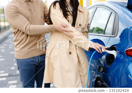 Young couple man and woman traveling by electric car having stop at charging station. Girlfriend holding power cable supply in hand while boyfriend gesturing finger up. 135102420