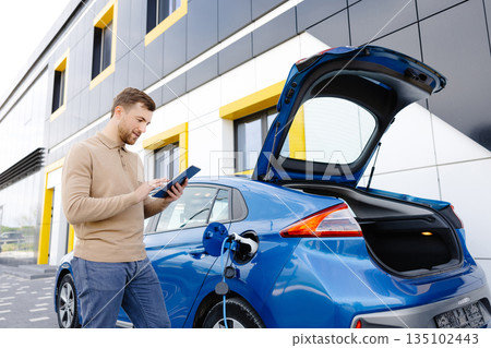 A young man is charging an electric car while standing at a station and holding a tablet A young man is charging an electric car while standing at a station and holding a tablet 135102443