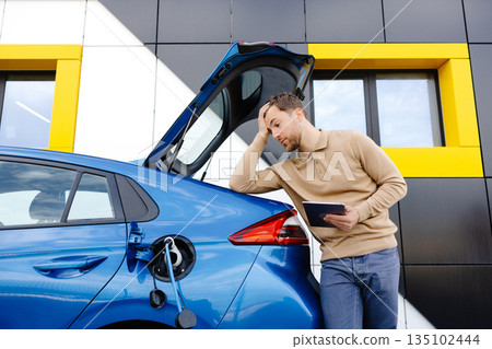 A young man is charging an electric car while standing at a station and holding a tablet 135102444