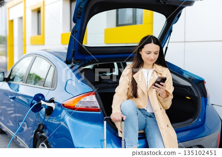 Young woman waiting charging automobile battery from small public station and using smartphone while charging automobile 135102451