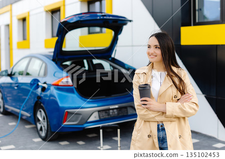 Young woman with cup of coffee waiting while her electric car charging, sustainable and economic transportation concept 135102453