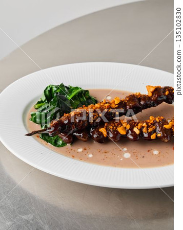 Photograph emphasizing glazed meat skewer and wilted vegetable garnishes Photograph emphasizing glazed meat skewer and wilted vegetable garnishes 135102830