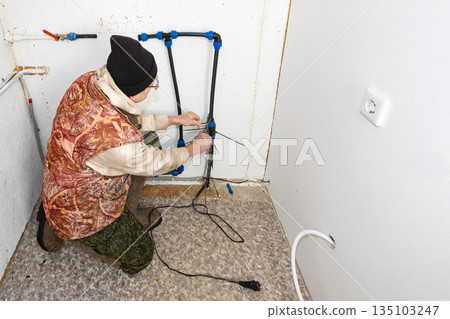 After disconnecting country house from water supply man installs heating cable on incoming pipe to prevent rupture during winter frosts. 135103247