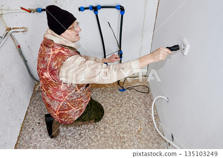 Man inserts electrical plug of heating cable for water pipe into socket in cold utility room with incoming water pipe. 135103249