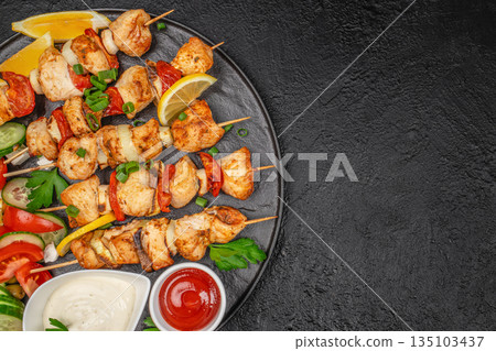 Grilled chicken skewers with vegetables, lemon and sauces on black plate over dark background, top view Grilled chicken skewers with vegetables, lemon and sauces on black plate over dark background, top view 135103437