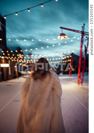 Woman walks on snowy street with string lights during evening hours in a festive setting 135103545
