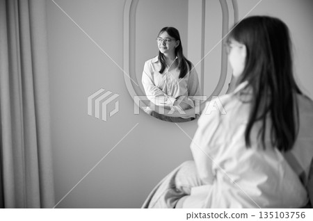 Beautiful girl looking at herself in mirror and smiling. Pretty female checking herself. Cute girl sitting in chair opposite to mirror at home. Black and white image. Beautiful girl looking at herself in mirror and smiling. Pretty female checking herself. Cute girl sitting in chair opposite to mirror at home. Black and white image. 135103756