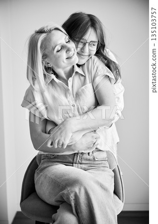 Cute family moment of mom and daughter hugging. Close up portrait of teenage girl showing her love to mother. Two female in casual clothes hugging with closed eyes at home. Black and white image. Cute family moment of mom and daughter hugging. Close up portrait of teenage girl showing her love to mother. Two female in casual clothes hugging with closed eyes at home. Black and white image. 135103757