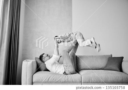 Joyful father lifting happy little son at home. Carefree father and son weekend. Happy young father lying on couch and fooling around with kid. Black and white image. Joyful father lifting happy little son at home. Carefree father and son weekend. Happy young father lying on couch and fooling around with kid. Black and white image. 135103758