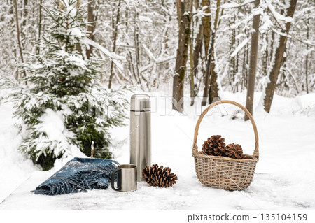 Picnic arrangement with thermos, stainless steel mug, pine cones and basket on snow table in winter forest. Cozy outdoor winter leisure and warm drink concept. 135104159