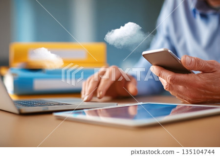 Hands using smartphone and tablet while working at a desk in a modern office with a laptop and cloud computing elements Hands using smartphone and tablet while working at a desk in a modern office with a laptop and cloud computing elements 135104744
