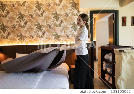 Young woman in hotel uniform doing bed in a hotel room Young woman in hotel uniform doing bed in a hotel room 135105032