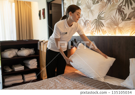 Young woman in hotel uniform doing bed in a hotel room Young woman in hotel uniform doing bed in a hotel room 135105034