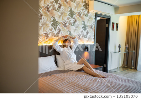 Woman in white bathrobe sitting on the bed in a hotel suit and looking relaxed 135105070