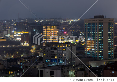 Night cityscape of Pristina, capital of Kosovo, showing modern high rise buildings, residential neighborhoods, illuminated windows, urban density and foggy winter atmosphere 135105253