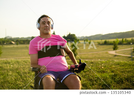 Wheelchair user enjoying music surrounded by green grass and sunlight. Wheelchair user enjoying music surrounded by green grass and sunlight. 135105340