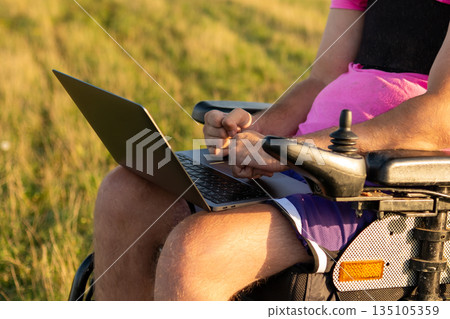 Man in wheelchair works on modern laptop holding device on lap in sunny meadow. Male freelancer enjoying business work via Internet in grassy meadow Man in wheelchair works on modern laptop holding device on lap in sunny meadow. Male freelancer enjoying business work via Internet in grassy meadow 135105359