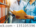 A healthcare professional assists a doctor while holding medical files. Soft daylight illuminates the room, highlighting the organized files and teamwork involved. 135105396