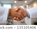 Two individuals engage in a handshake inside a consultation room, signifying a professional interaction during a medical appointment. 135105406