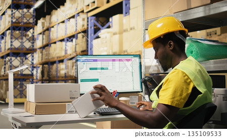 Black depot clerk reviewing inventory data on tracking software for distribution system, busy logistics hub stock room with organized racks. Online retail fulfillment and supply chain. Camera A. Black depot clerk reviewing inventory data on tracking software for distribution system, busy logistics hub stock room with organized racks. Online retail fulfillment and supply chain. Camera A. 135109533
