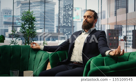 CEO practices mindfulness in his modern office listening to meditation music through a mobile app. Calm posture and closed eyes reflect a moment of peace amid corporate pressures. Camera A. CEO practices mindfulness in his modern office listening to meditation music through a mobile app. Calm posture and closed eyes reflect a moment of peace amid corporate pressures. Camera A. 135109545