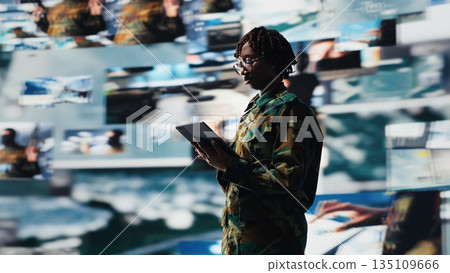 Military officer in war room oversees mission operations using tablet app. African american army soldier using device application to do surveillance and threat monitoring, camera B 135109666
