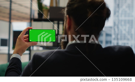 Focused chief executive officer looks at chroma key display on phone, examining infographics data in his office. Young business administrator implementing investment strategy. Camera B. 135109668