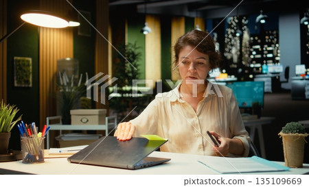 Female employee checking the time on her phone and closing laptop, preparing to leave office late at night after doing overtime. Intense workload makes her stay after hours. Camera A. 135109669