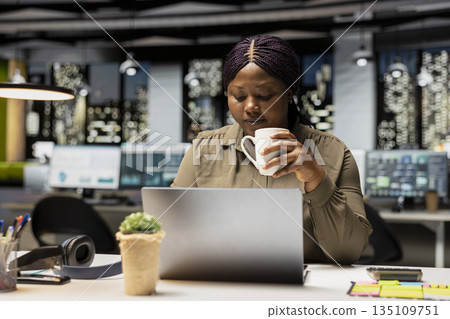 Black woman specialist enjoying a cup of coffee to stay awake late at night, handling administrative tasks in the corporate space. Proofreading and writing at night with a caffeine drink. Black woman specialist enjoying a cup of coffee to stay awake late at night, handling administrative tasks in the corporate space. Proofreading and writing at night with a caffeine drink. 135109751