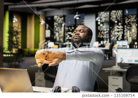Black male consultant in discomfort due to neck stiffness and backache, stretching at desk to relieve the pain and strain. Businessman struggling with incorrect posture at the office after hours. Black male consultant in discomfort due to neck stiffness and backache, stretching at desk to relieve the pain and strain. Businessman struggling with incorrect posture at the office after hours. 135109754