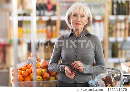 Portrait of mature woman shopper with shopping cart choosing some products in supermarket Portrait of mature woman shopper with shopping cart choosing some products in supermarket 135111076