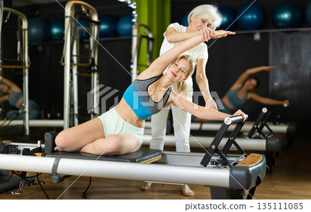 Senior woman trainer helps young woman to perform exercises in sitting position on pilates reformer Senior woman trainer helps young woman to perform exercises in sitting position on pilates reformer 135111085