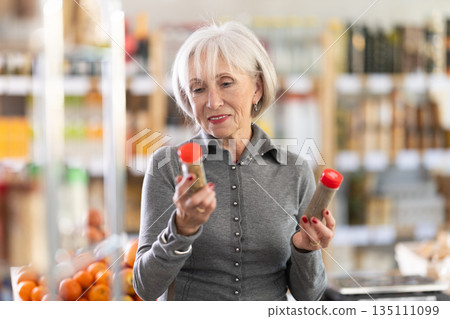 Elderly woman chooses spices and seasonings to buy in grocery department section of store Elderly woman chooses spices and seasonings to buy in grocery department section of store 135111099